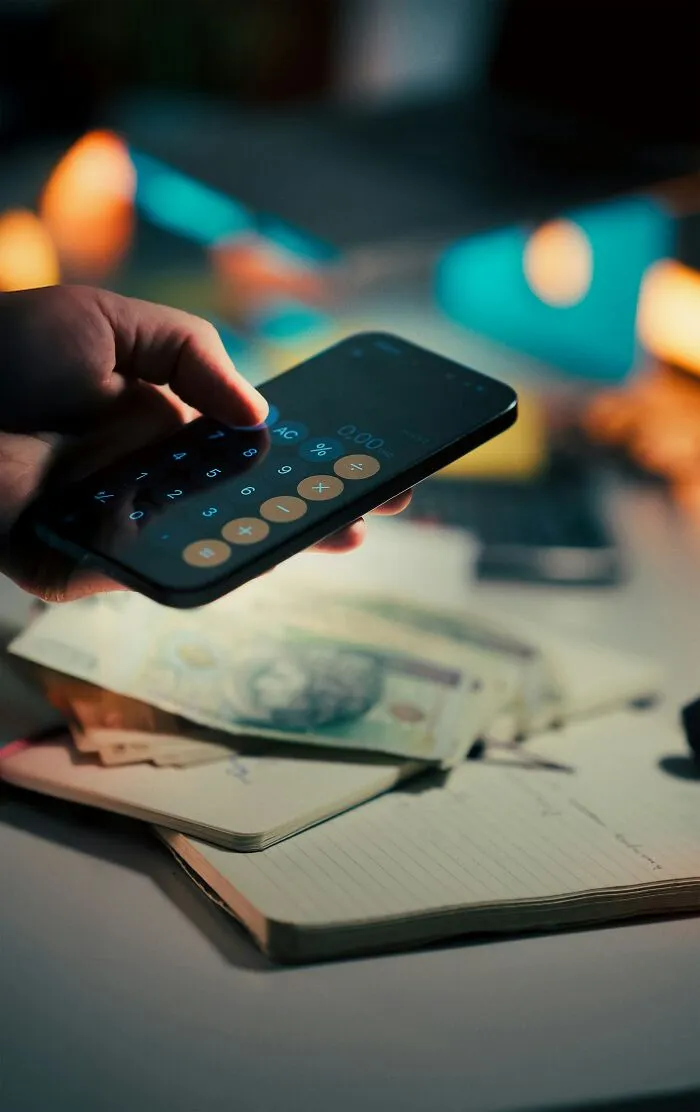 A hand holding a smartphone showing a calculator app over a stack of money, highlighting cheap habits.