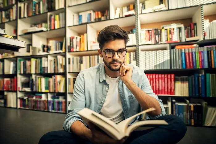 Young man reading a book in a library, reflecting on his career options in a difficult job market.