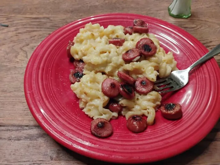 Macaroni and cheese topped with burnt sliced hot dogs on a red plate, showing classic food combinations.