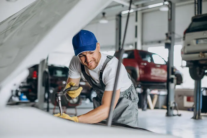A male auto mechanic smiling while working on a car engine, depicting a job where women face subtle sexism.