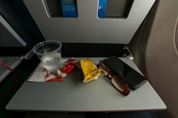An airplane tray table with a cup of water, snacks, glasses, and a phone, showing a plane seatmate setup.