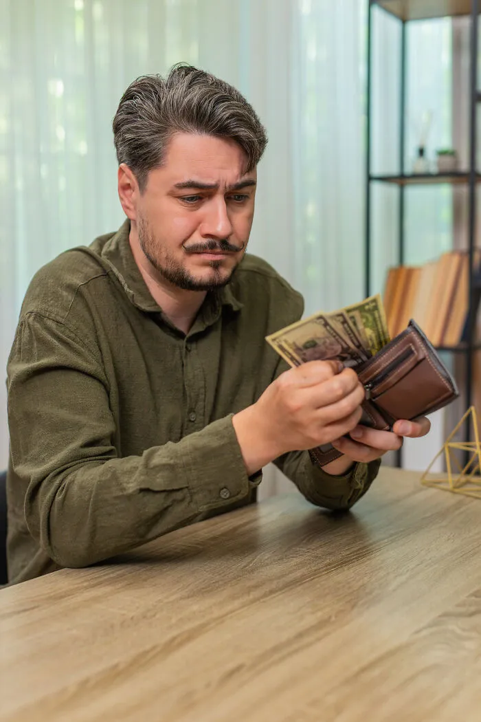 A worried man looking into his leather wallet with only a few dollars left, demonstrating cheap living.