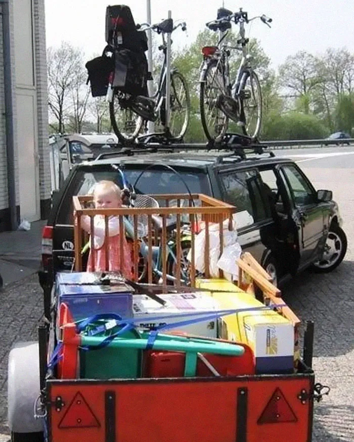 Child in crib and household items loaded on trailer behind car with bikes on roof, a moving fails setup.