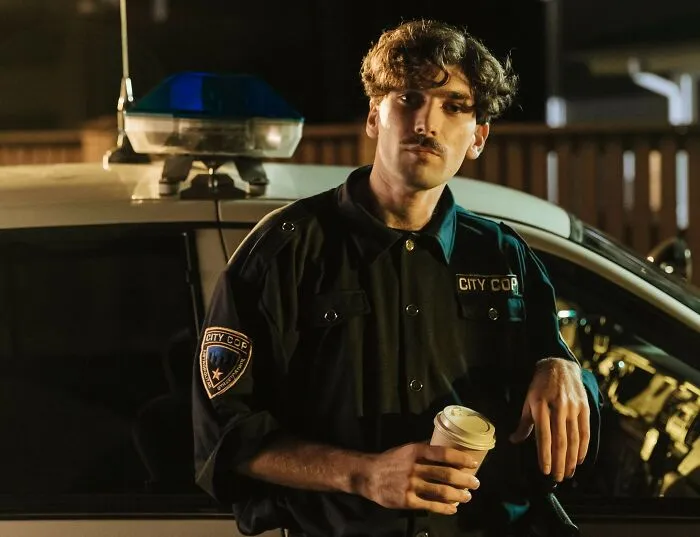 City cop in uniform holding coffee cup beside police car at night
