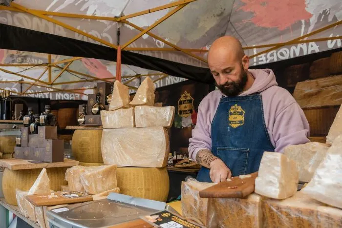 A cheesemonger slicing cheese at an outdoor market stall, highlighting middle-class life preferences.