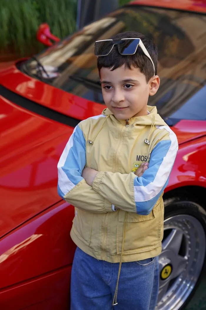 A young boy posing with crossed arms next to a red car, symbolizing the dreams of a middle-class life.