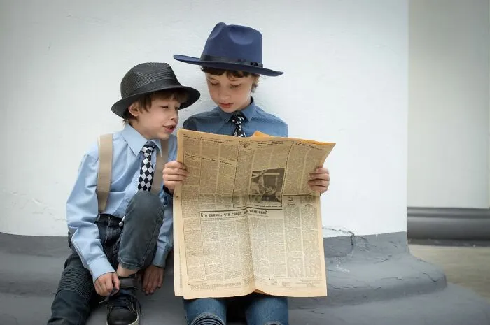 Two young boys wearing fedoras and reading a newspaper, portraying middle-class life.