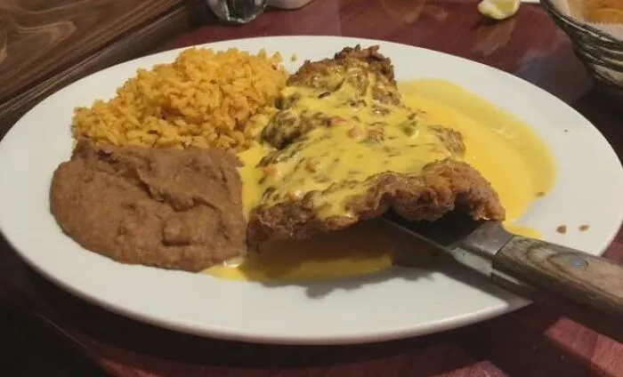 Unique food combinations featuring a fried cutlet covered in yellow cheese sauce next to rice and refried beans