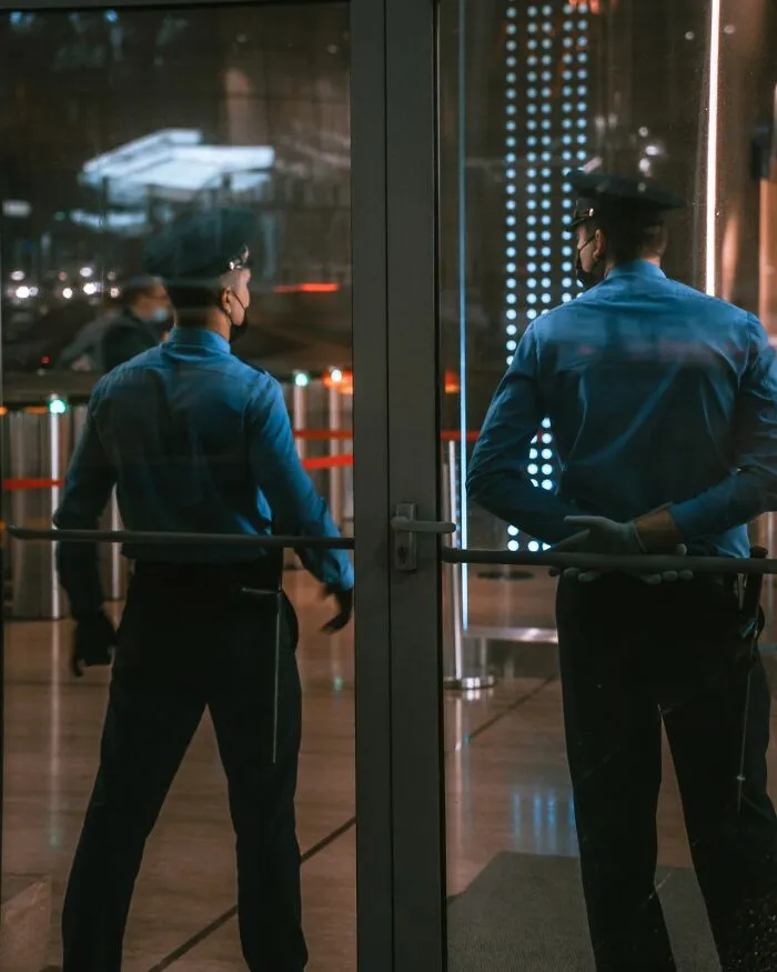 Two service workers, security guards, seen from behind, reflected in glass, wearing blue uniforms and masks.