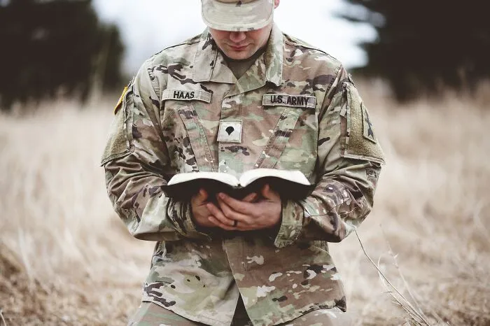 A US Army soldier kneeling and reading a book in a field, representing a life path of service for smart kids.