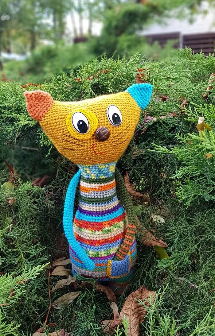 A yellow crochet cat toy with striped body stands nestled in green evergreen bushes.