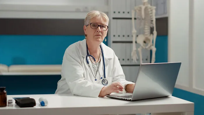 A female doctor wearing glasses and a lab coat looking at the camera while using a laptop.