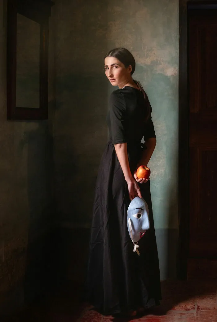 A dramatic portrait photo of a woman in a black dress holding a blue mask and a red apple behind her back