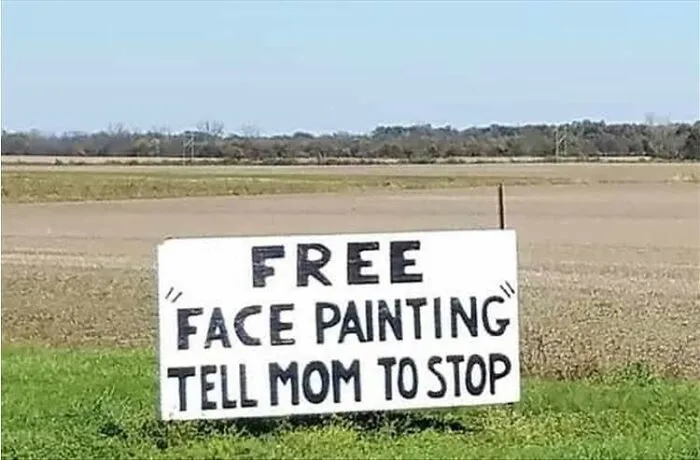 A roadside sign in a field offering free face painting, featuring misused quotation marks around the service name.