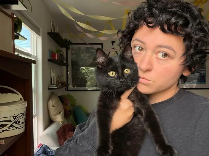 A person with dark curly hair posing next to a fluffy black cat, showing pets that look like their owners.