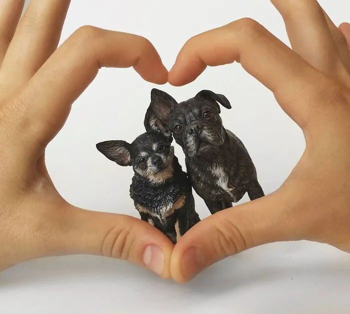 Hands making a heart shape around two small hyperrealistic sculptures of pets, both tiny black dogs.