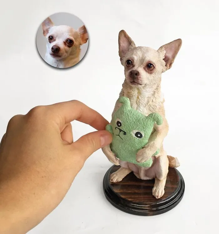 A human hand placing a toy in a hyperrealistic sculpture of a chihuahua sitting on a wooden stand