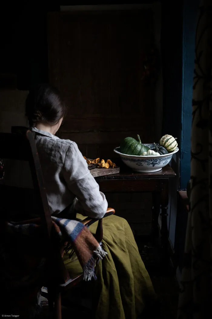 A person seated near a rustic table featuring a bowl of small pumpkins