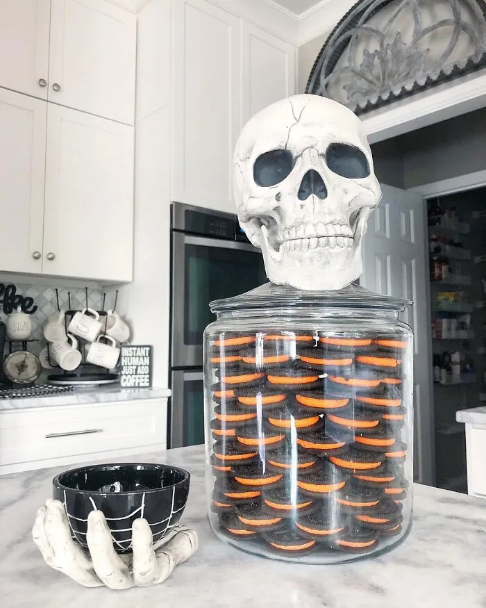Halloween skull decoration sitting atop a large glass jar filled with black and orange cookies.