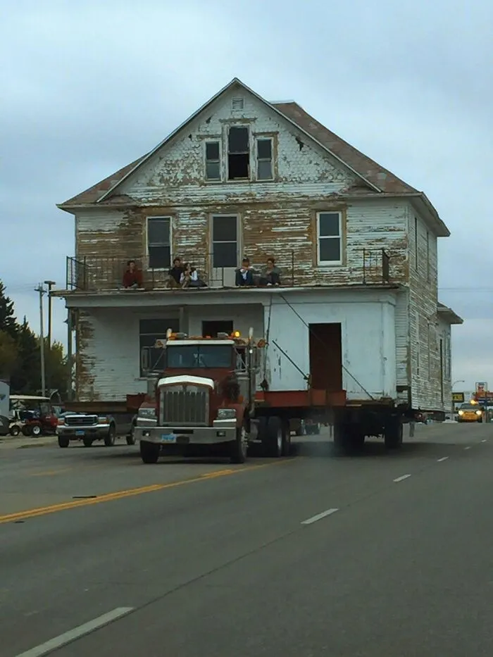 A large wooden house being transported on a truck down a road, creating cool and entertaining pics