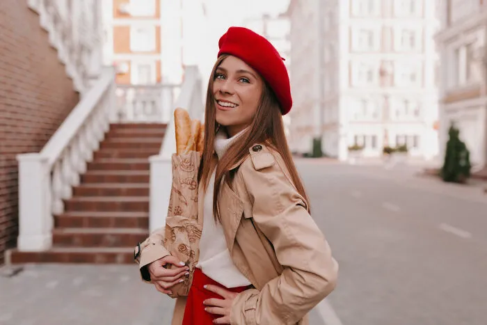 A stylish woman wearing a red beret holding french bread, looking like an obvious lie.