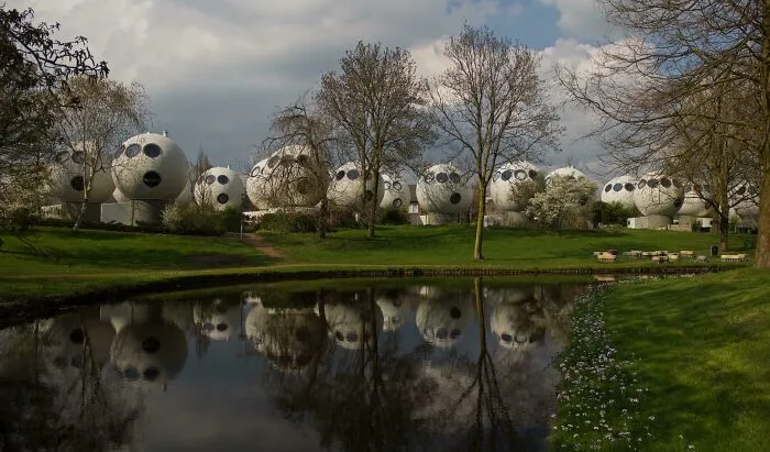 A row of white, spherical buildings showing bizarre shapes on a grassy bank near a pond.