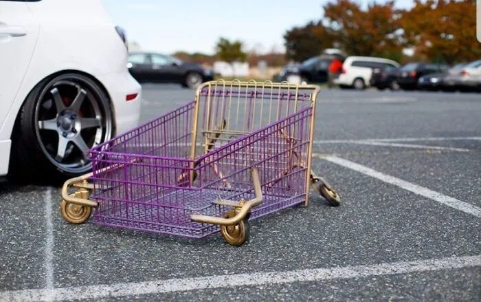 A purple shopping cart modified into a lowered vehicle sitting in a parking lot next to a sports car.