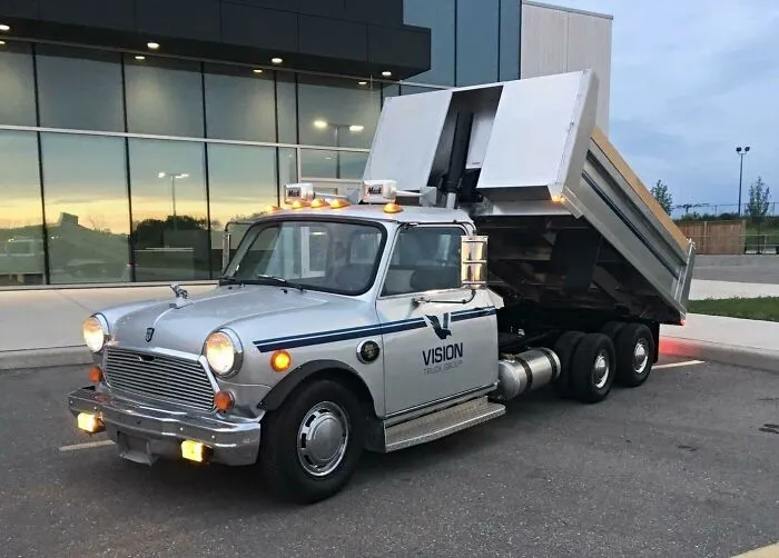 A unique silver dump truck featuring a classic car front cab design parked outside a modern glass building.