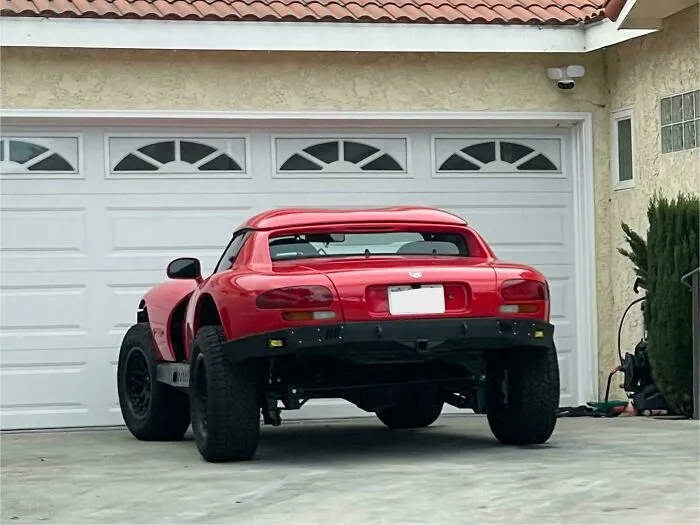 Rear view of a modified red sports car with oversized off-road tires parked in front of a residential garage.