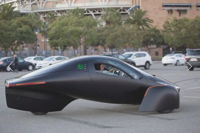 A futuristic black three-wheeled electric vehicle driving through a urban parking lot.