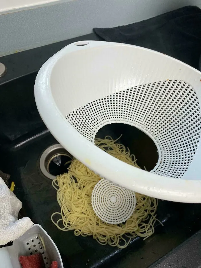 Cooked spaghetti spilled directly into a kitchen sink through a broken plastic colander.