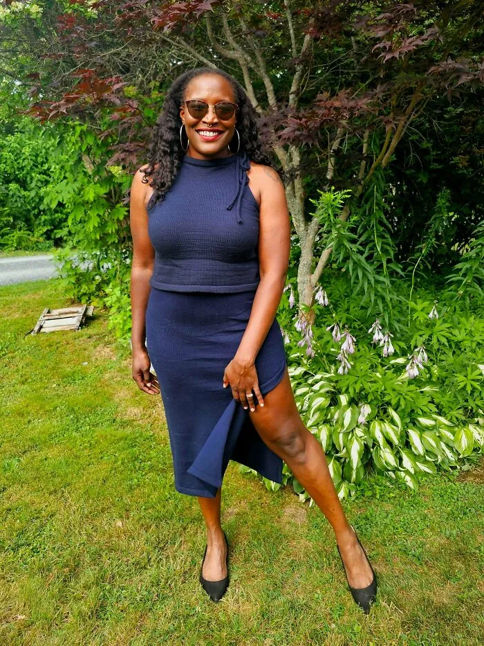 A smiling woman wearing a hand-knit blue top and matching slit skirt, showcasing creative knitting.