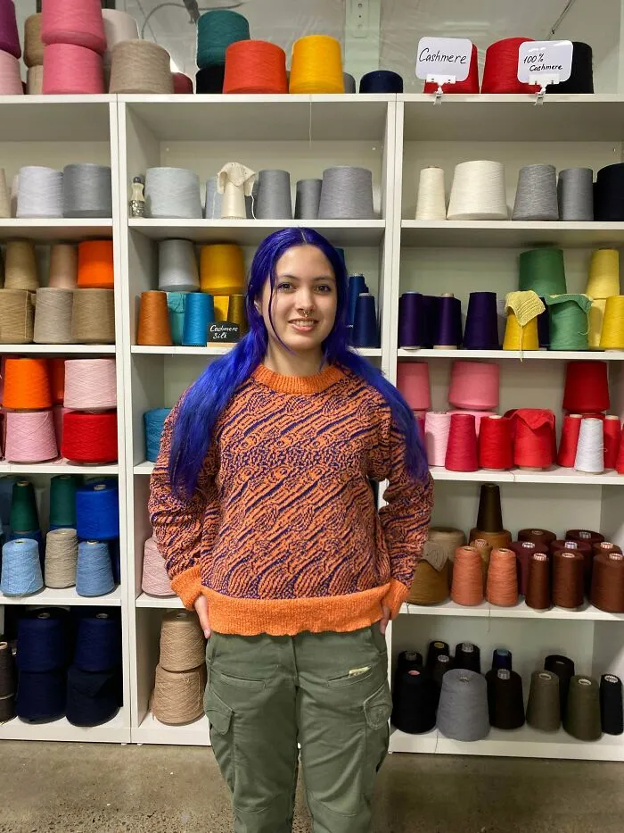A girl with purple hair posing in front of yarn shelves wearing a knitted sweater