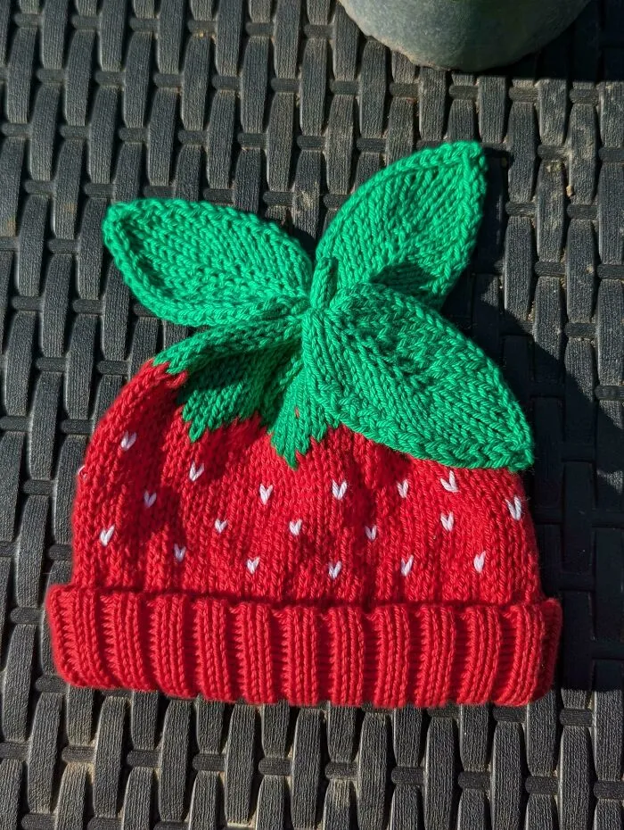 A red knitted hat shaped like a strawberry with green leaves on top, created by knitting pros