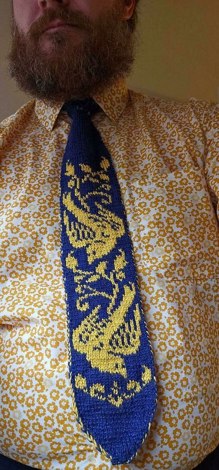 A man wearing a blue and yellow knitted tie with bird motifs, showing knitting skills.