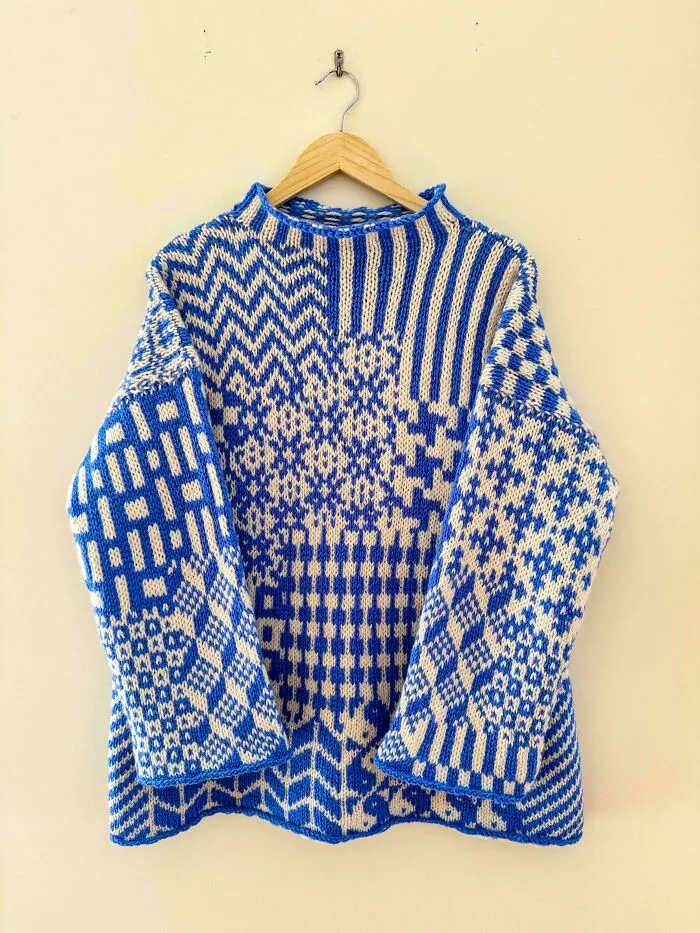 A blue and white geometric patterned knitted sweater hanging on a wall, showcasing advanced knitting