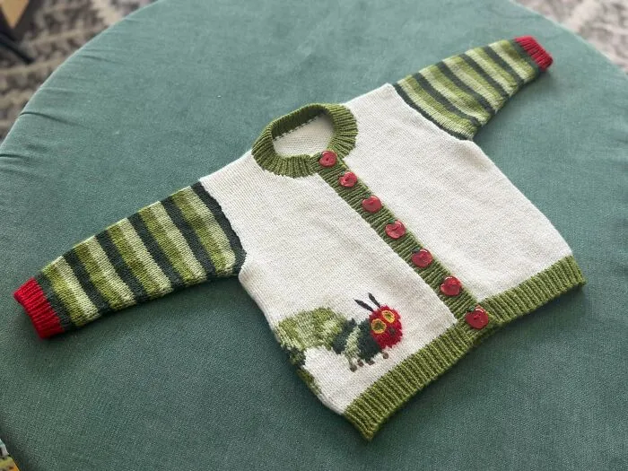 A white and green baby cardigan with a knitted Hungry Caterpillar design, showcasing creative knitting ideas