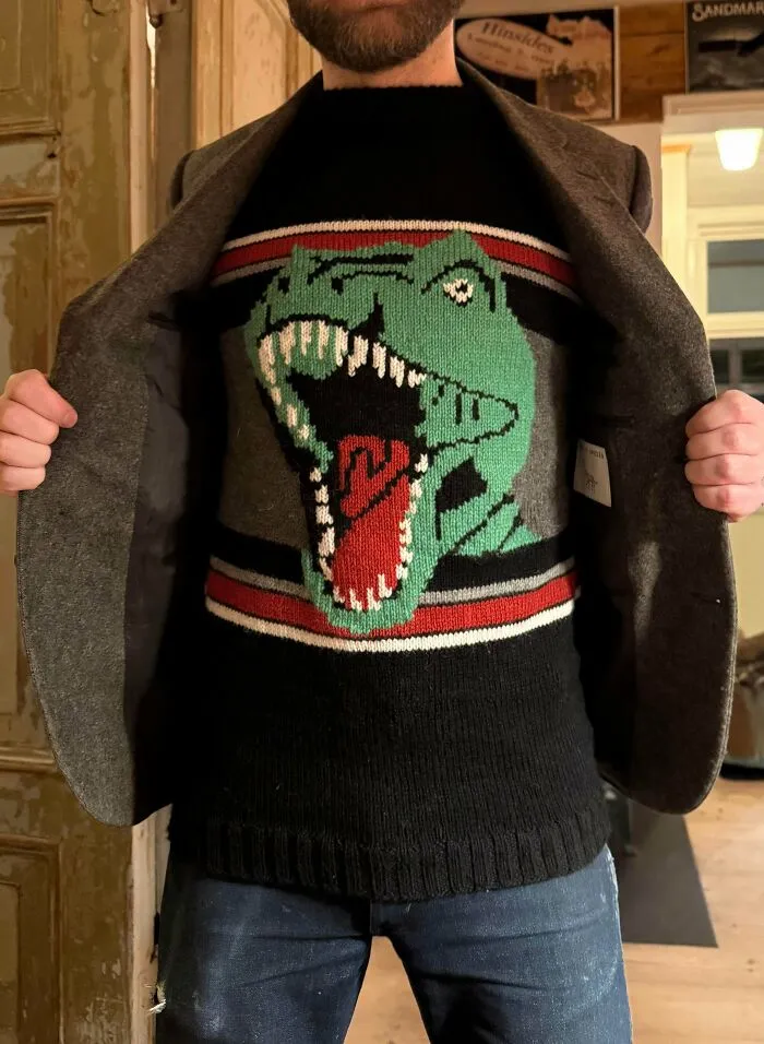 A man wearing a black knitted sweater with a green T-Rex design under a blazer, displaying knitting ideas
