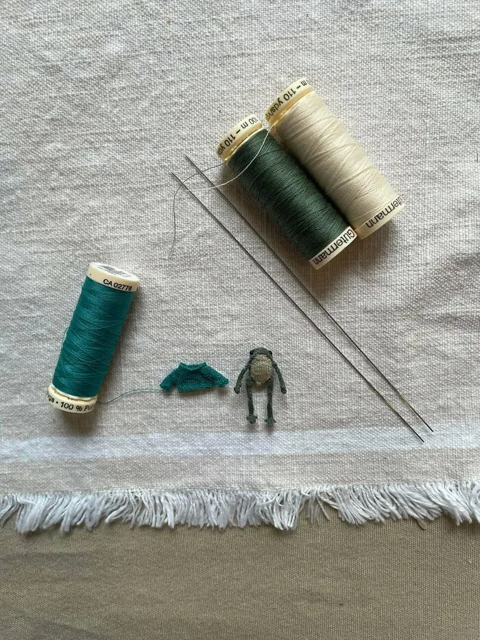 A tiny green frog toy and miniature sweater next to spools of thread made by knitting amateurs and pros