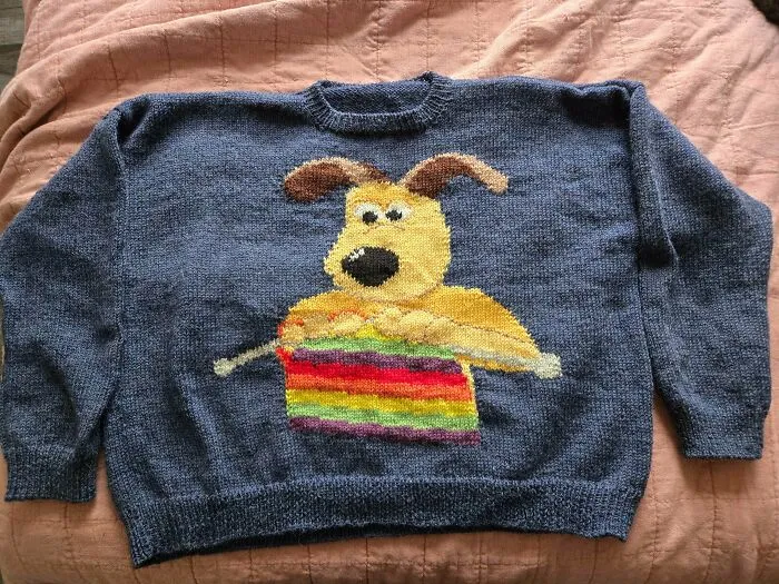 A blue sweater featuring a knitted dog knitting a colorful scarf by knitting amateurs and pros