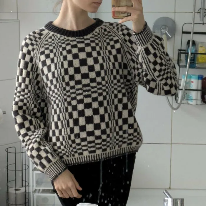 A person wears a black and white checkered optical illusion knitting sweater in a mirror selfie.