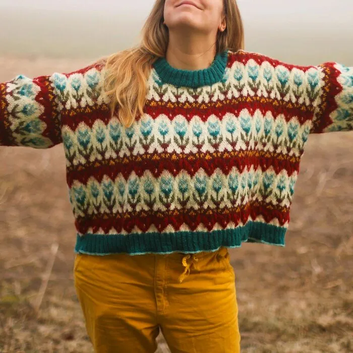 A woman wearing a vibrant, detailed knitting sweater with a red and blue floral pattern outdoors.