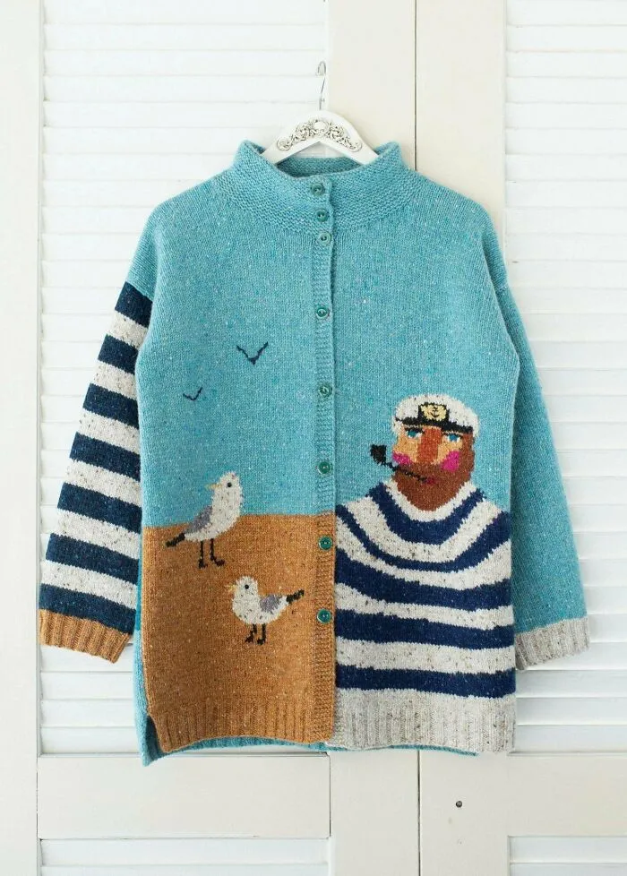 A detailed blue cardigan showcasing knitting pros work with a hand-knit sailor and seagulls beach scene