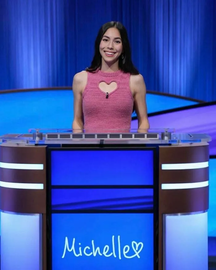 A young woman wearing a pink hand-knitted sleeveless top with a heart cutout on a TV game show set.