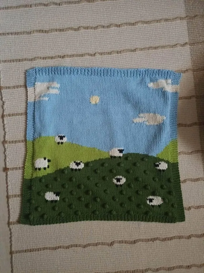 A hand-knitted wall hanging depicting a green field with white sheep under a blue sky, representing knitting amateurs.