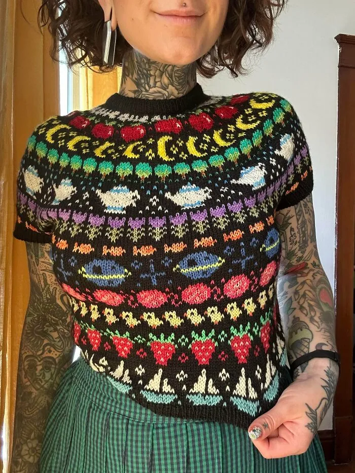 A woman wearing a black short-sleeved knitted top with colorful patterns, created by knitting amateurs and pros