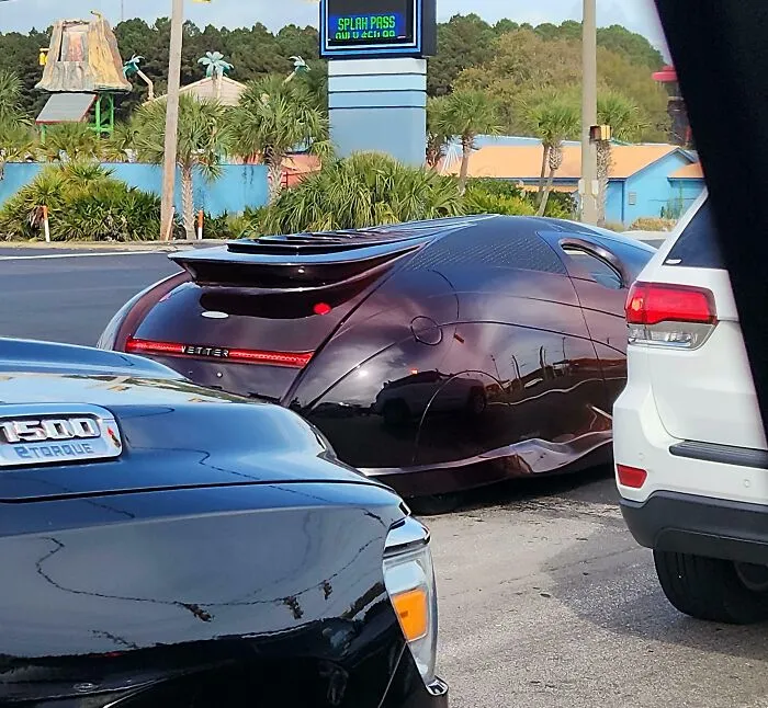 A dark purple custom futuristic sports car parked on a city street near other normal cars.