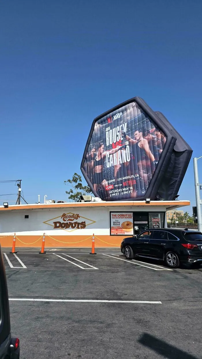 An inflatable billboard of a Netflix wrestling match on a Randy's Donuts building, illustrating modern life in America.