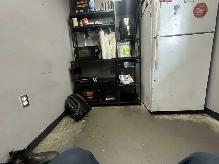 A cramped breakroom with basic appliances and a backpack on the floor, capturing an aspect of modern life in America.
