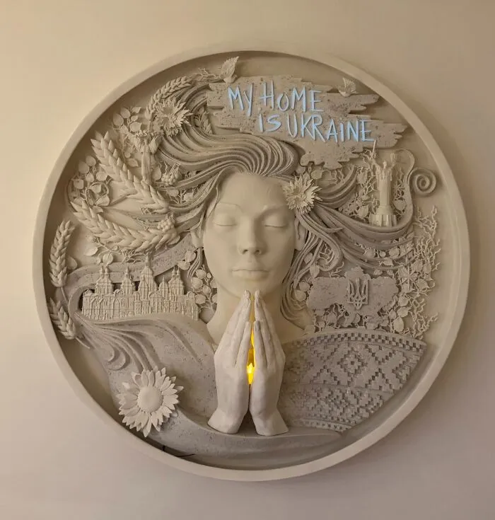 An intricate circular wall relief sculpture made from 3D prints depicting a praying woman and Ukrainian symbols.